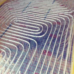 Białe rury ogrzewania podłogowego ułożone spiralnie na folii izolacyjnej z czerwoną siatką, przy ścianie zabezpieczonej żółtą taśmą.