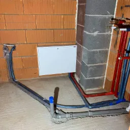 Surowe wnętrze z instalacją hydrauliczną i grzewczą w trakcie montażu: rury w izolacji (czerwone i niebieskie) prowadzone wzdłuż ściany z cegły i betonowego filaru, szare rury kanalizacyjne...