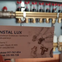 Rozdzielacz instalacji grzewczej w szafce z wizytówką firmy Instal Lux, specjalizującej się w instalacjach grzewczych i sanitarnych, z widocznymi danymi kontaktowymi.