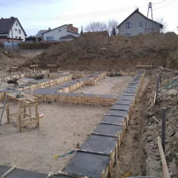 Wykop pod fundamenty z drewnianym szalunkiem wypełnionym świeżym betonem, widoczny wózek budowlany i stół roboczy z narzędziami, w tle ziemia i domy.