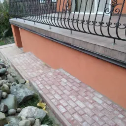 Ceglana ścieżka z kostki brukowej prowadząca do pomalowanej na pomarańczowo elewacji budynku z kutą, czarną balustradą i kamieniami dekoracyjnymi po lewej stronie.