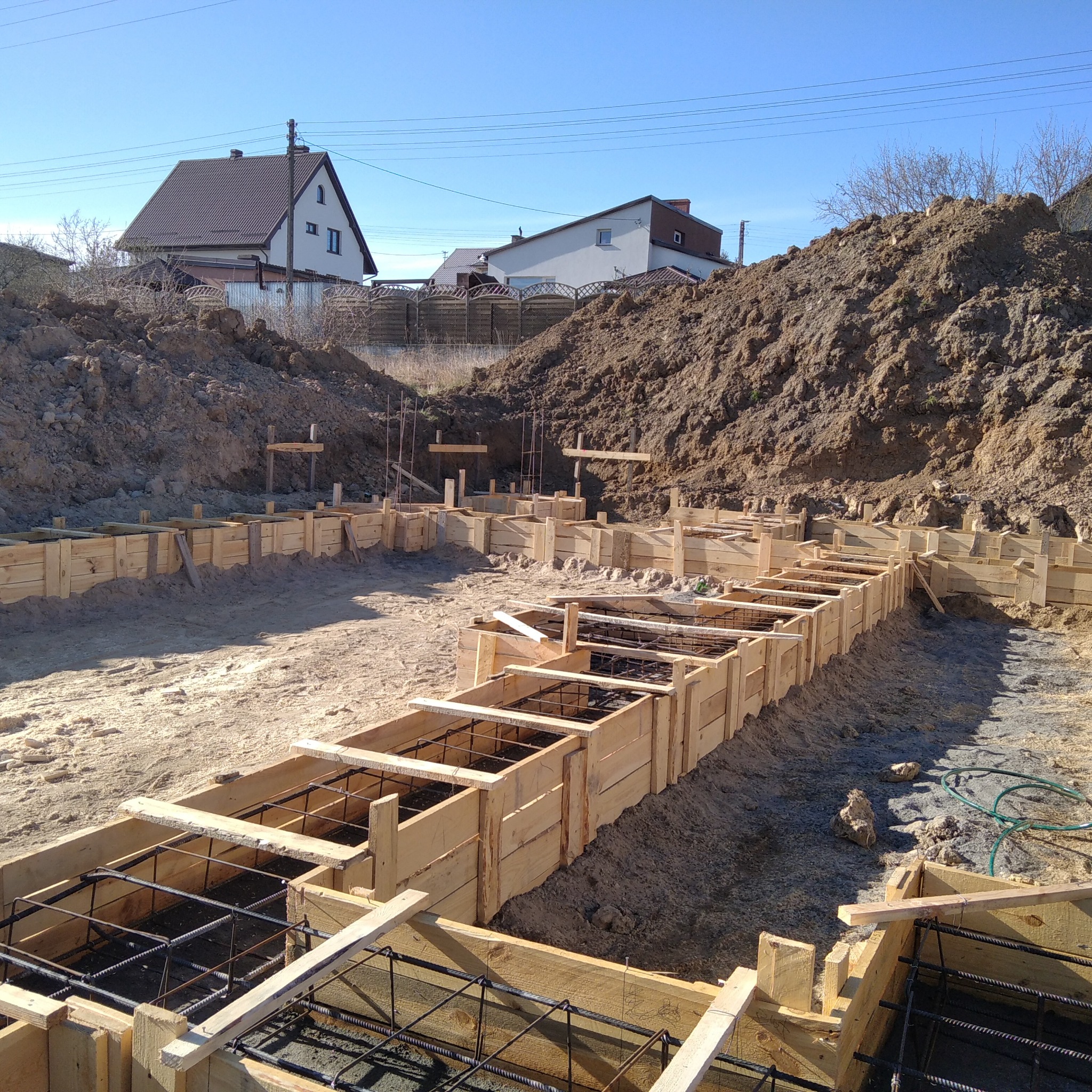 Drewniany szalunek zbrojenia fundamentów budynku na tle wykopu i domów jednorodzinnych w słoneczny dzień.