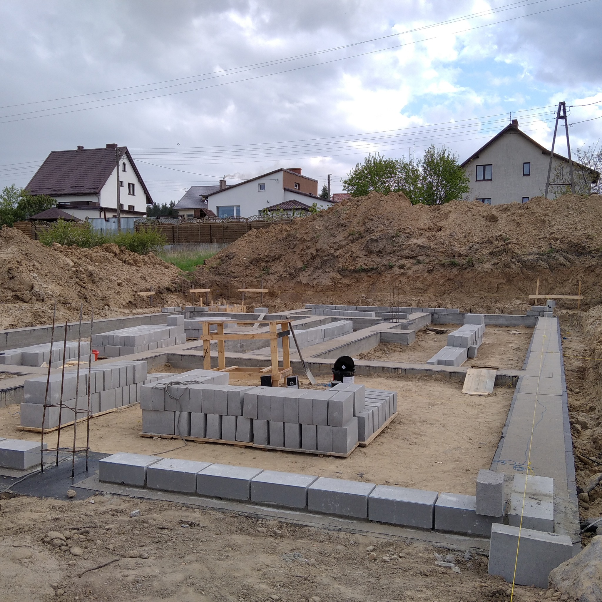 Budowa fundamentów domu jednorodzinnego: ułożone bloczki betonowe na zaprawie, widoczne szalunki, zbrojenie, ziemia dookoła i domy w tle.
