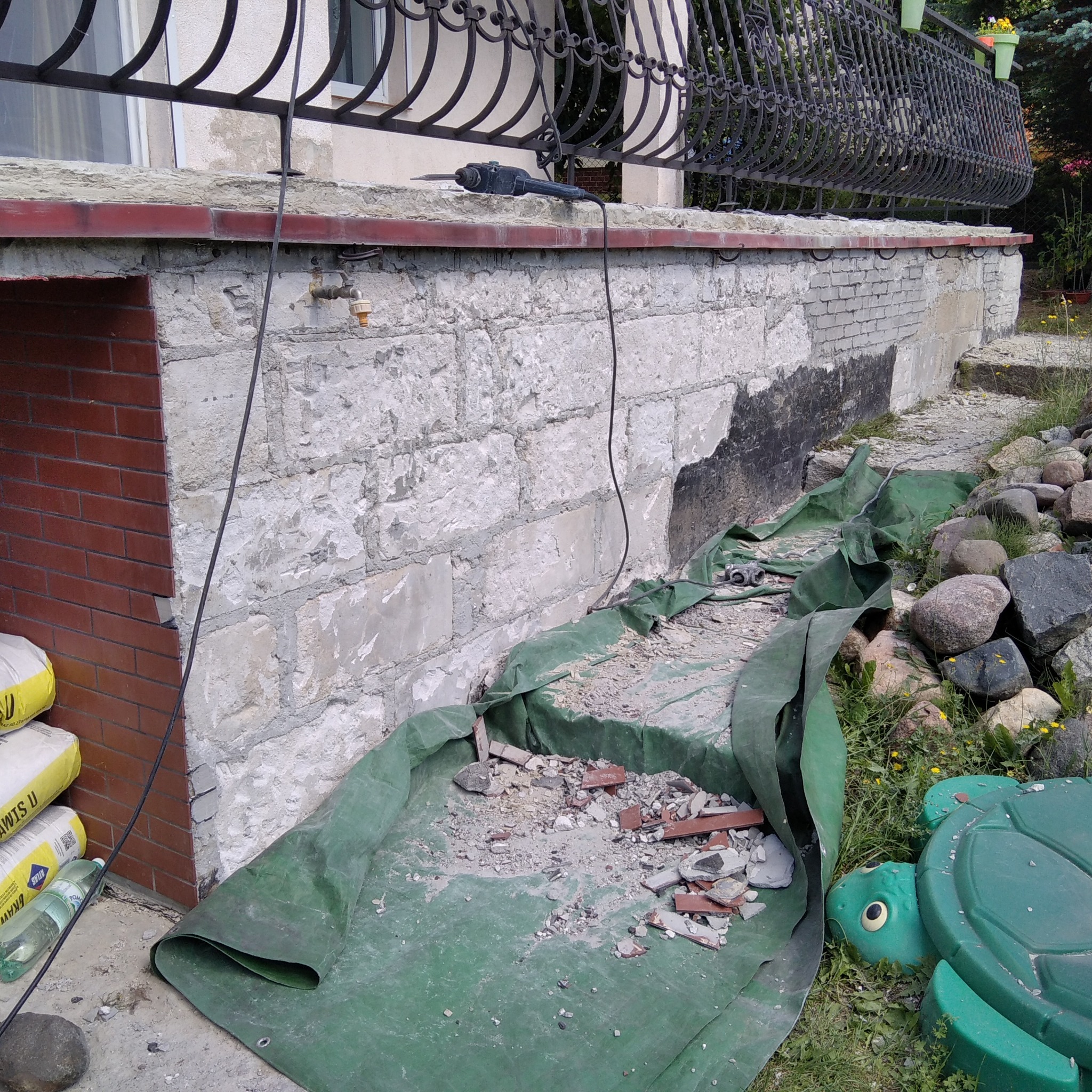 Demontaż starej elewacji z białej cegły na fundamencie domu, widoczny fragment nowej ceglanej obudowy, narzędzie do demontażu na górze, gruz zabezpieczony zieloną folią, worki z zaprawą...