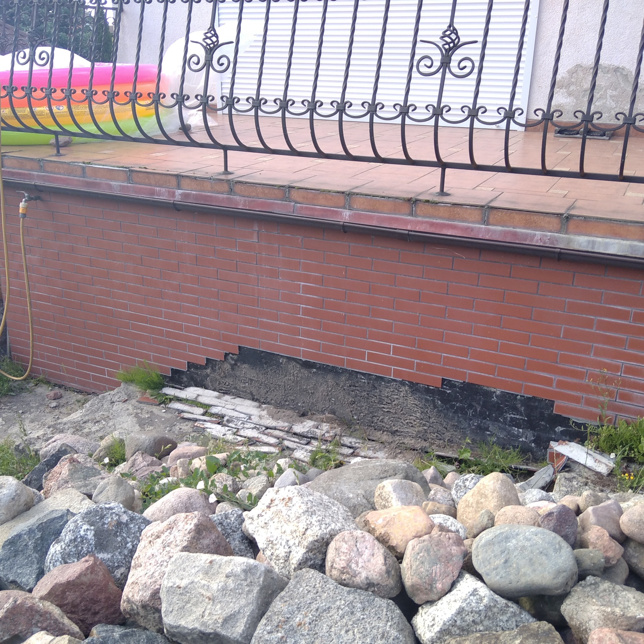 Ceglana elewacja fundamentu z częściowo usuniętą warstwą izolacyjną, odsłaniająca czarną hydroizolację, w tle widoczny fragment balustrady i rozłożony basen.