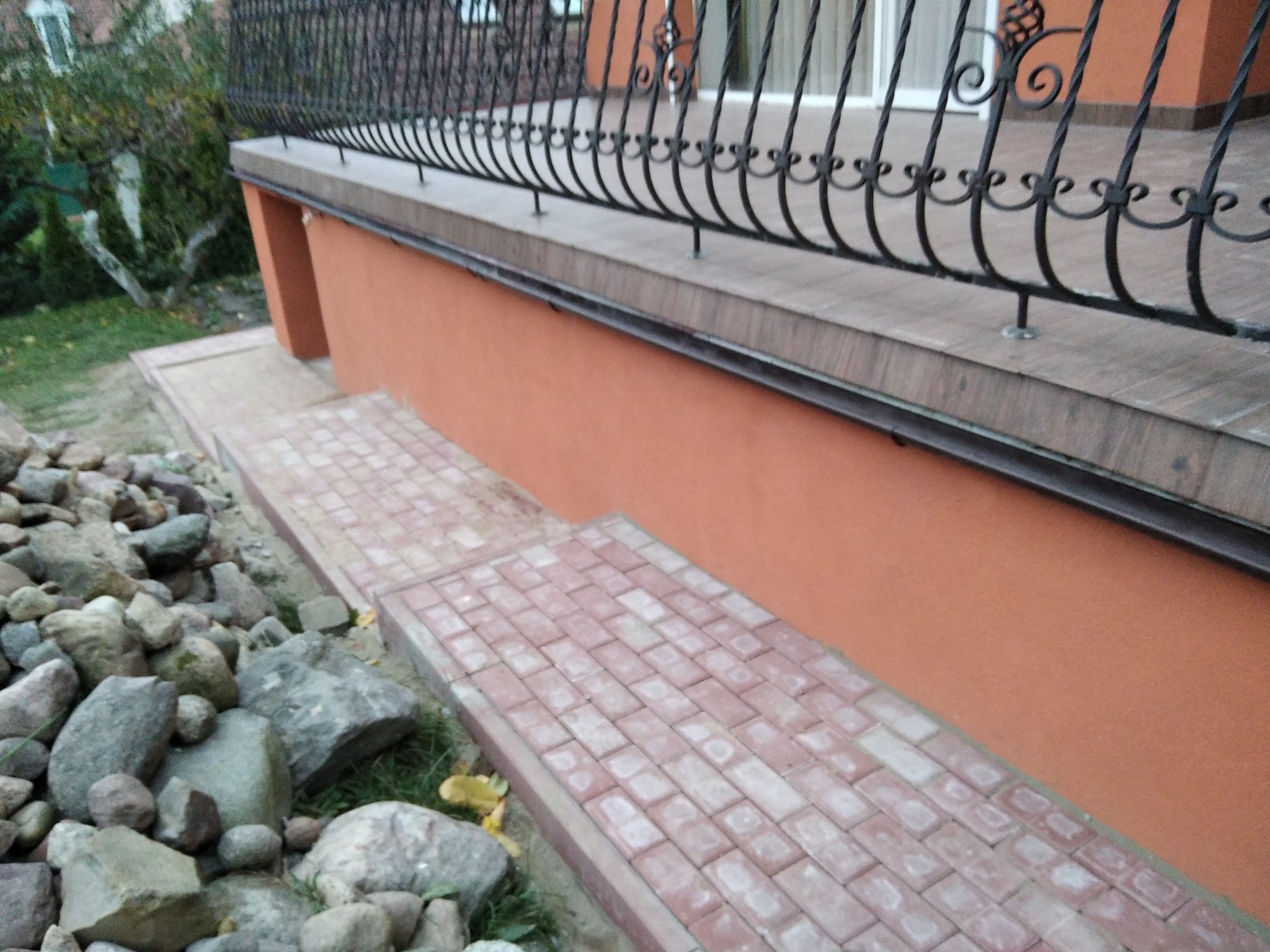 Ceglana ścieżka z kostki brukowej prowadząca do pomalowanej na pomarańczowo elewacji budynku z kutą, czarną balustradą i kamieniami dekoracyjnymi po lewej stronie.
