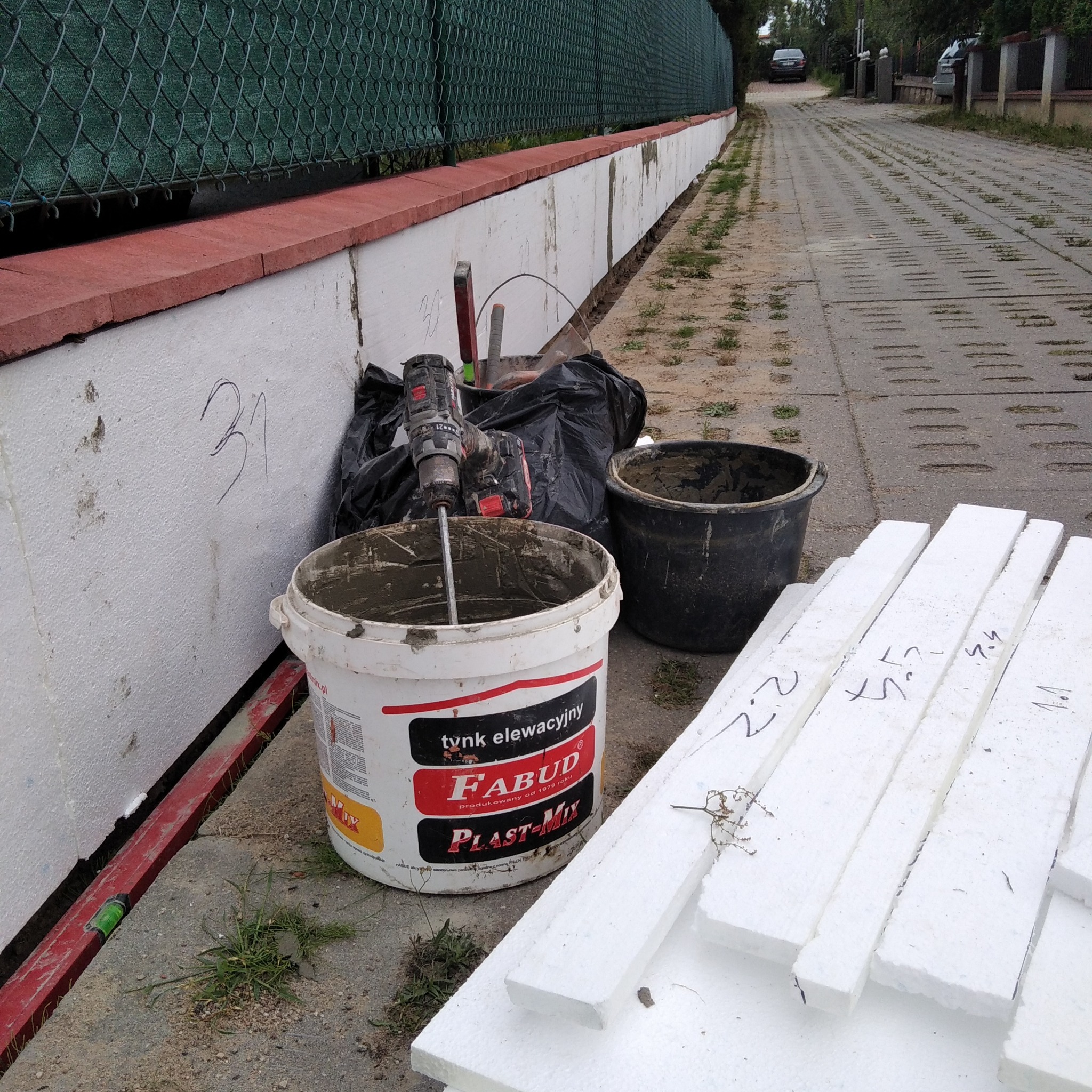 Przygotowanie do ocieplenia fundamentów: wiadro z zaprawą, mieszadło elektryczne, styropianowe płyty z naniesionymi oznaczeniami, czarne wiadro i worek na tle ocieplonego fragmentu fundamentu...