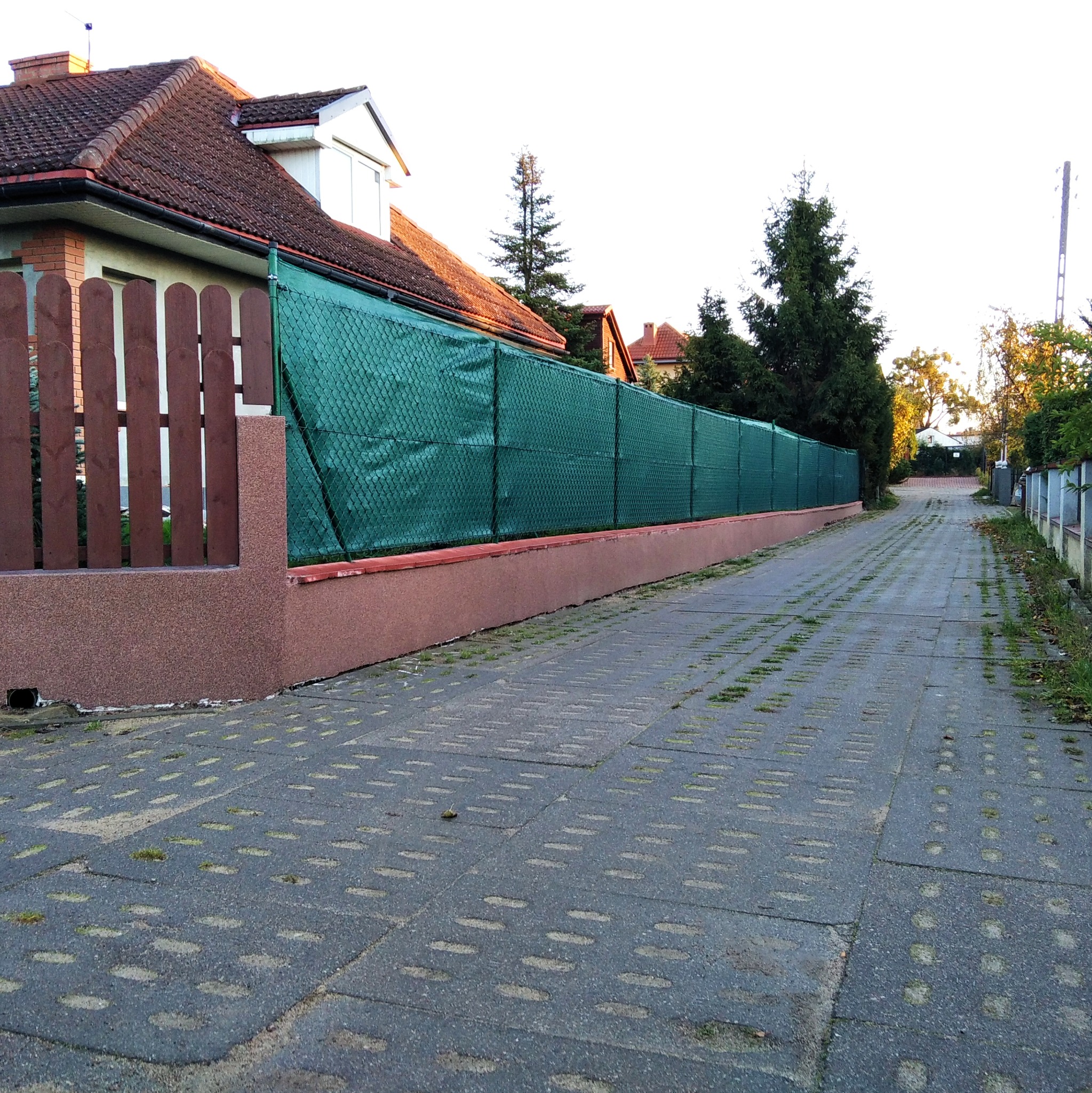 Zielona siatka maskująca na ogrodzeniu posesji z brązowym cokołem i drewnianymi sztachetami, widok z betonowej drogi z ażurowymi płytami.