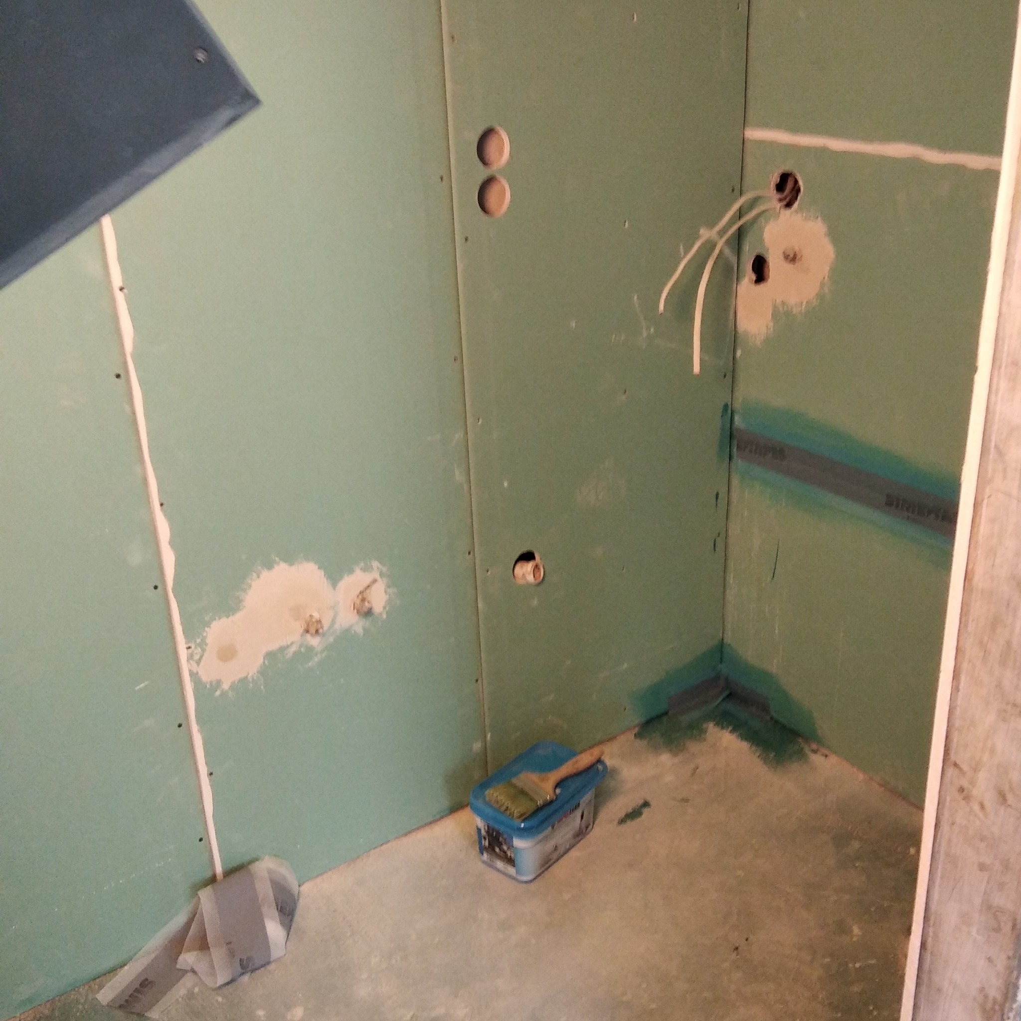 Łazienka w trakcie remontu: zielone płyty gipsowo-kartonowe zamontowane na ścianach, widoczne otwory na instalacje, zabezpieczone narożniki i szczeliny, puszka z farbą i pędzel na podłodze.