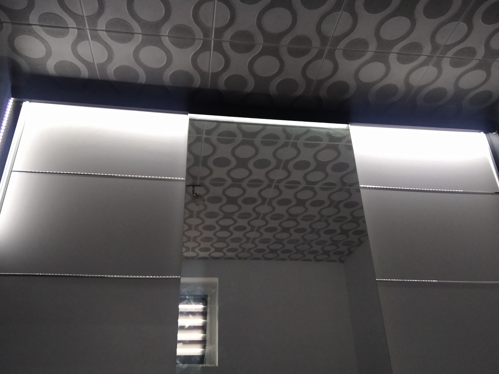 Nowoczesna łazienka z podświetlanym sufitem i lustrami, widoczne geometryczne płytki sufitowe i fragment okna z żaluzjami.