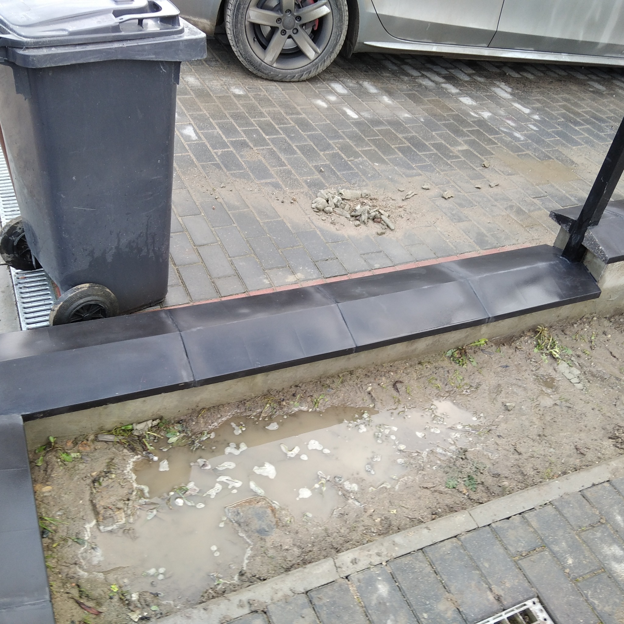 Ułożona czarna krawędź chodnika z widoczną studzienką kanalizacyjną, obok stojący czarny pojemnik na śmieci i fragment samochodu.