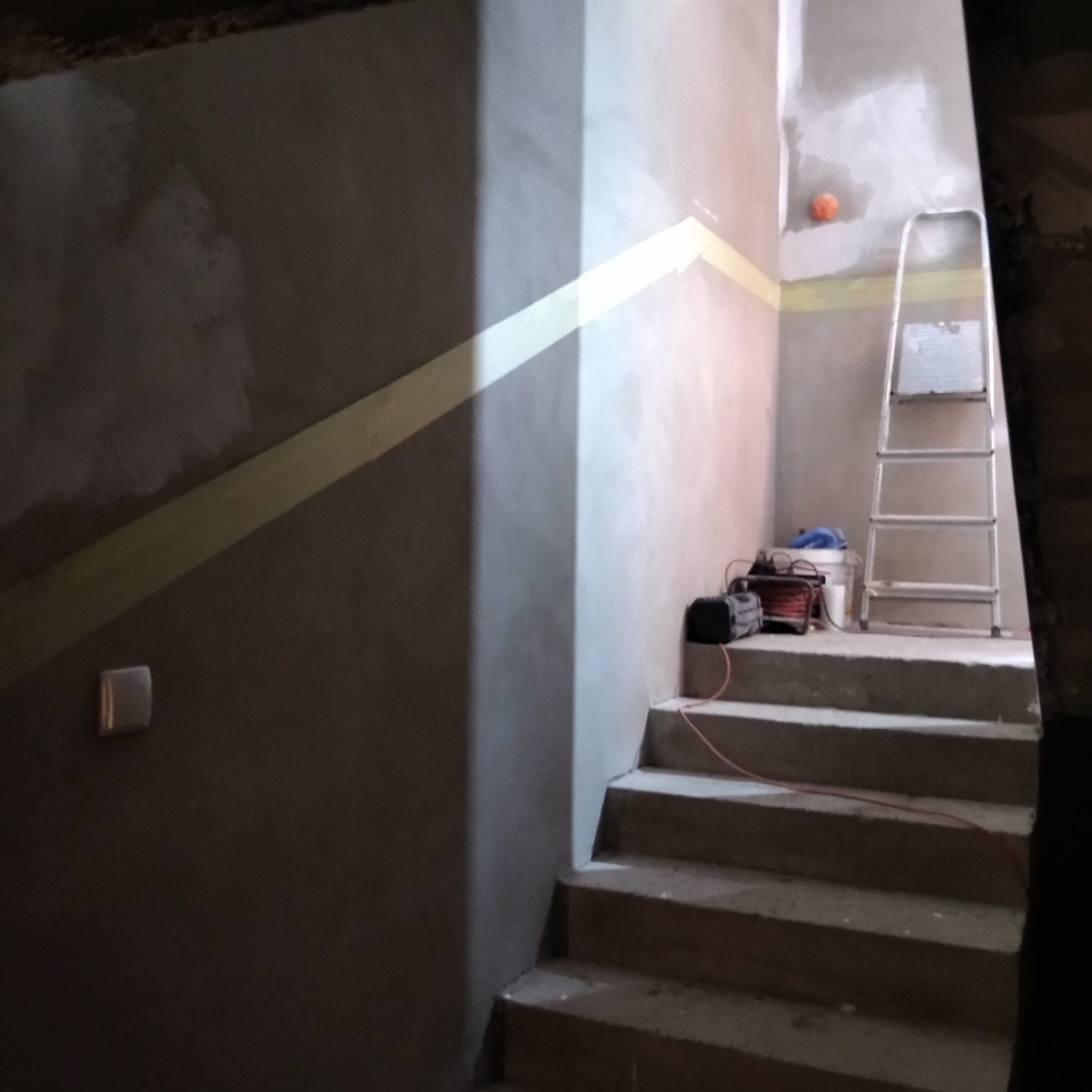 Klatka schodowa w trakcie prac wykończeniowych, ściany z świeżo nałożonym tynkiem, zabezpieczone taśmą malarską, drabina aluminiowa, wiadro i narzędzia na schodach, widoczny włącznik światła...