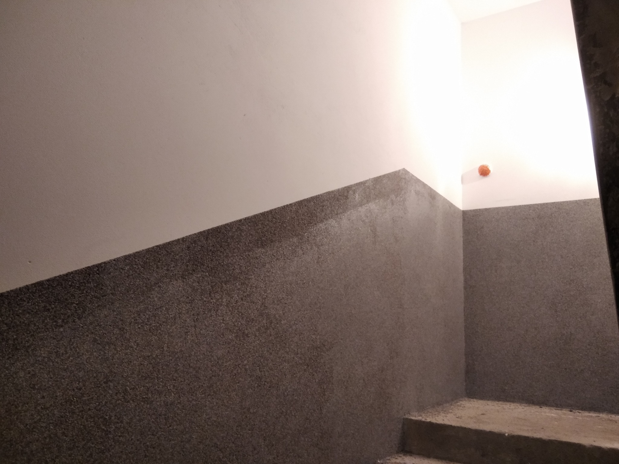 Dwuwarstwowa ściana klatki schodowej: górna część pomalowana na biało, dolna pokryta szarym, ziarnistym tynkiem strukturalnym, widoczny fragment schodów i pomarańczowa zaślepka po instalacji.
