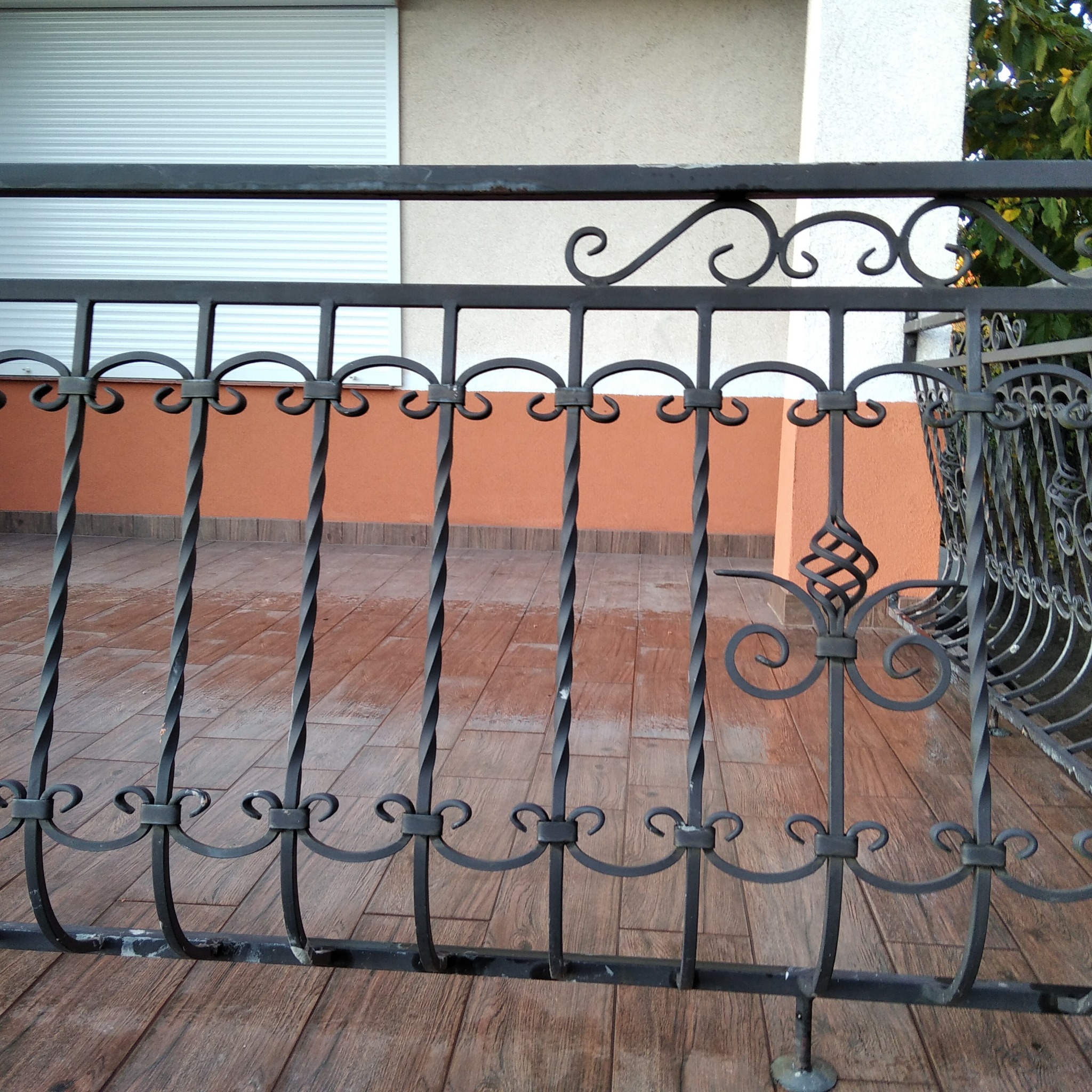 Czarna, metalowa balustrada balkonowa z dekoracyjnymi elementami, widok na pomarańczową elewację z białymi płytkami i częściowo zasłonięte rolety.