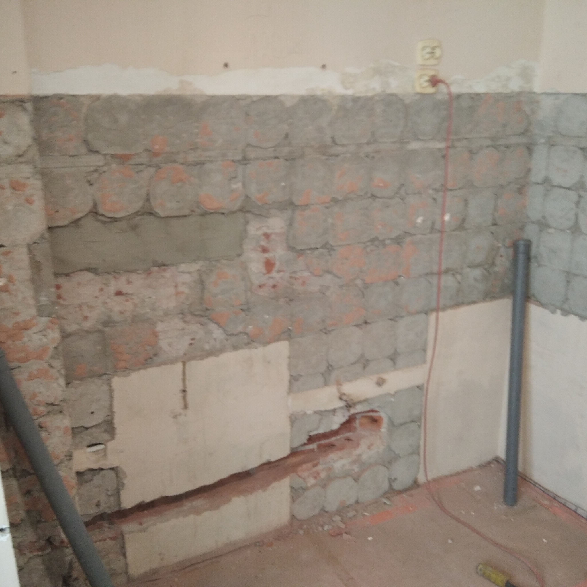 Ściana w trakcie remontu łazienki z odsłoniętą cegłą i fragmentami starej glazury, widoczne rury instalacyjne i gniazdko elektryczne.