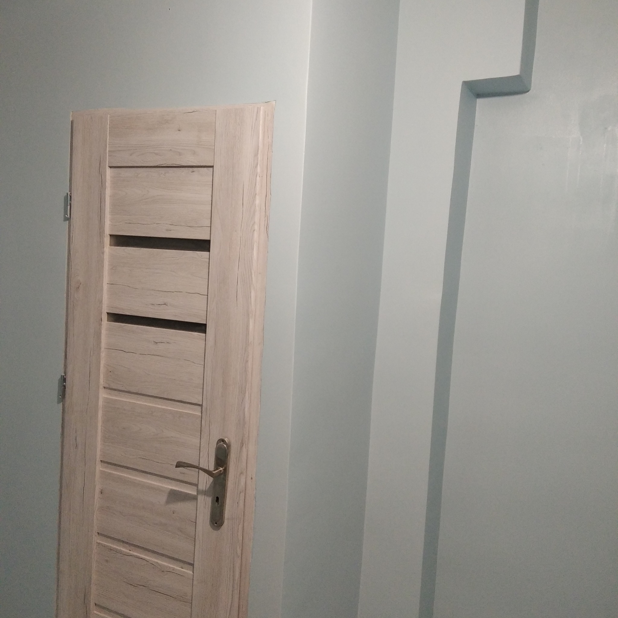 Zamknięte drzwi wewnętrzne z klamką, w odcieniach jasnego drewna, na tle pomalowanej na jasno szaro-niebiesko ściany z dekoracyjnym, wystającym elementem.