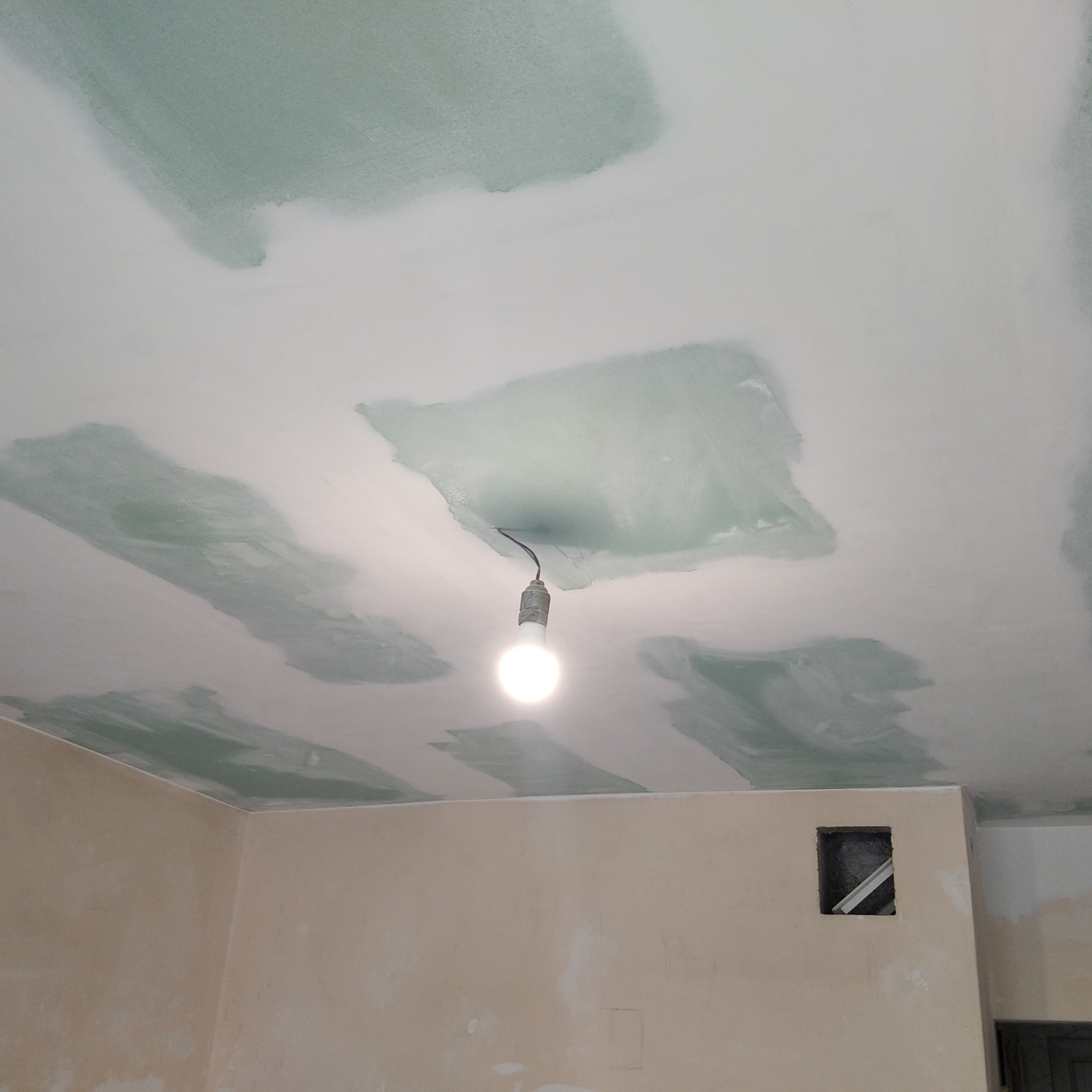 Sufit w trakcie szpachlowania z widocznymi smugami świeżo nałożonej gładzi, oświetlony wiszącą żarówką na kablu, ściany w jasnym odcieniu z widocznymi śladami po pracach remontowych i otworem...