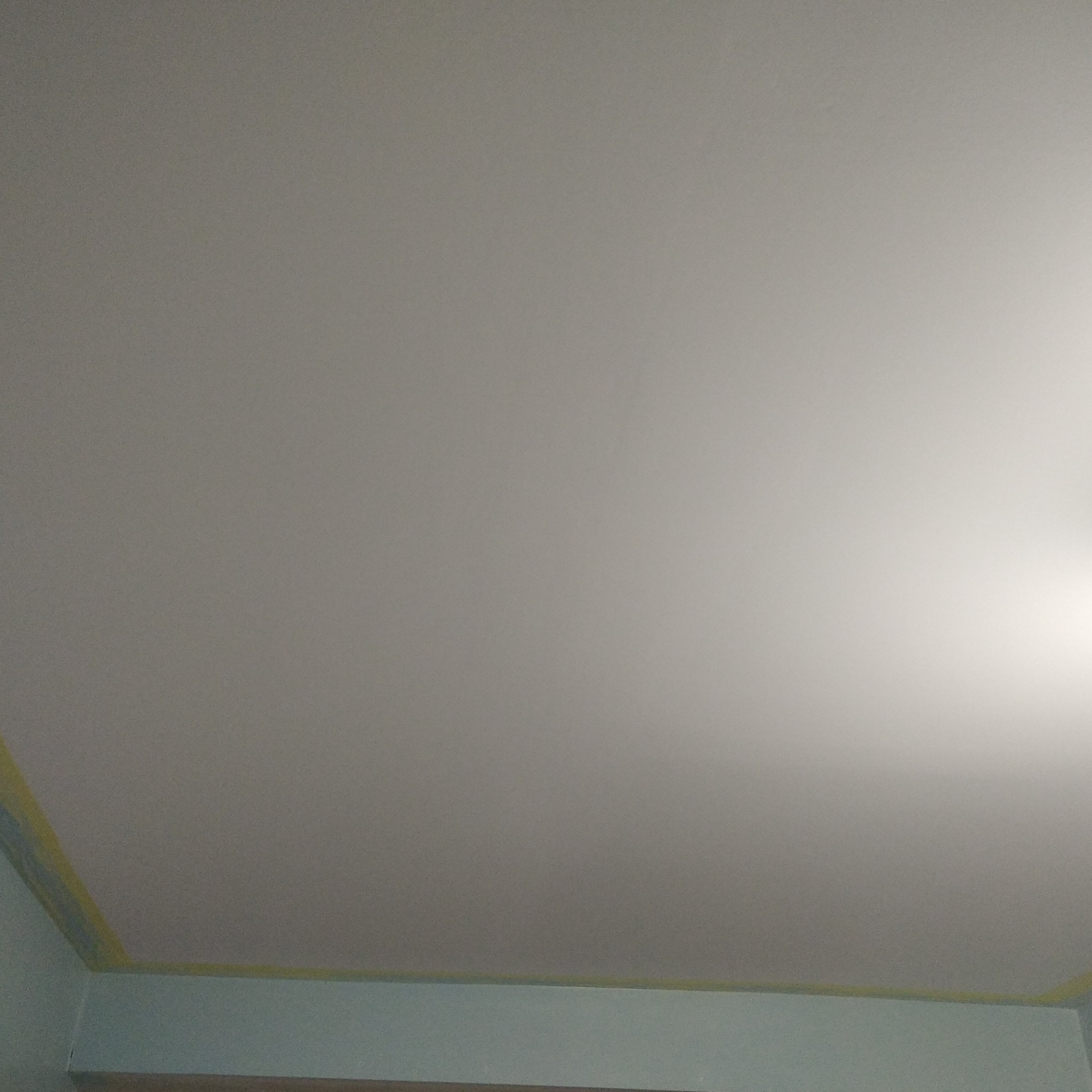 Świeżo pomalowany sufit w pomieszczeniu, zabezpieczony taśmą malarską przy ścianach, z widocznym fragmentem ściany w odcieniach błękitu.