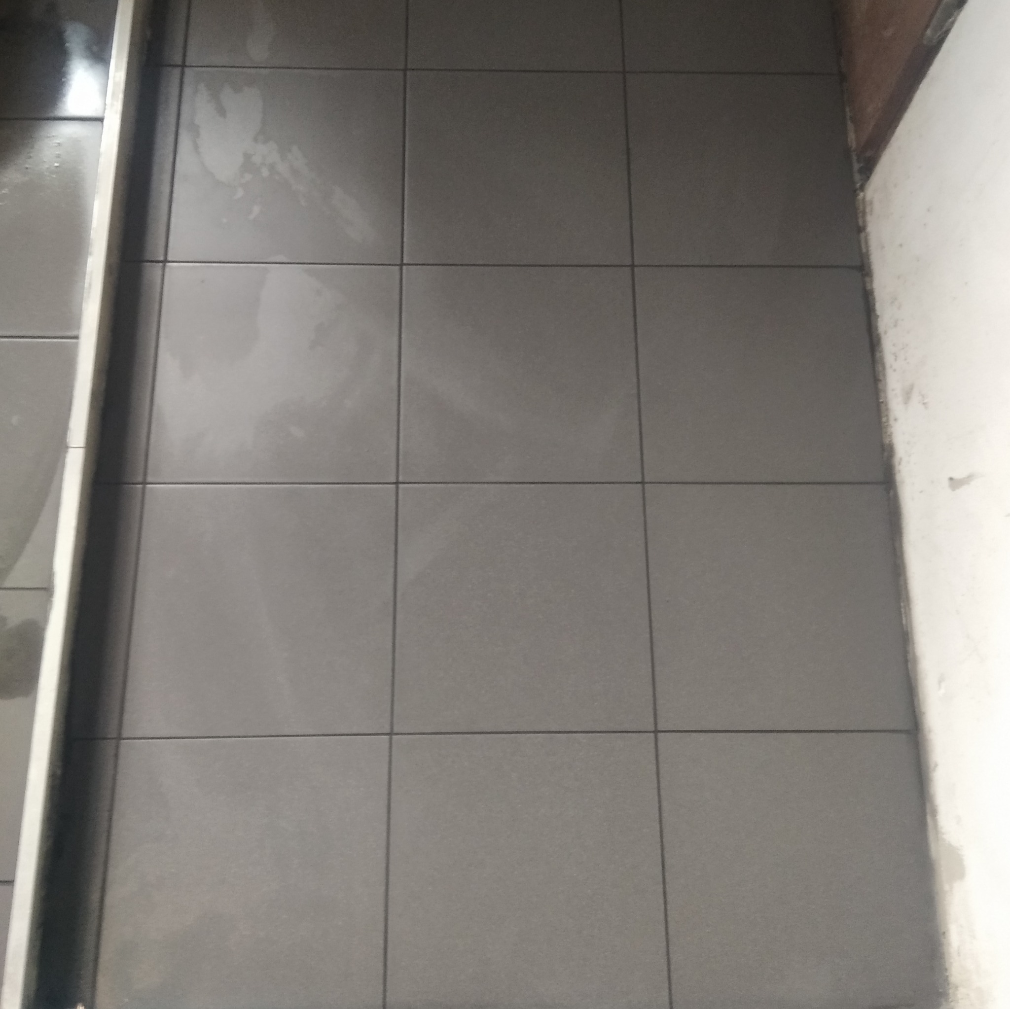 Świeżo ułożona, szara, kwadratowa podłoga z płytek w łazience, widoczne ślady po fugowaniu i czyszczeniu, przy krawędzi ściany.