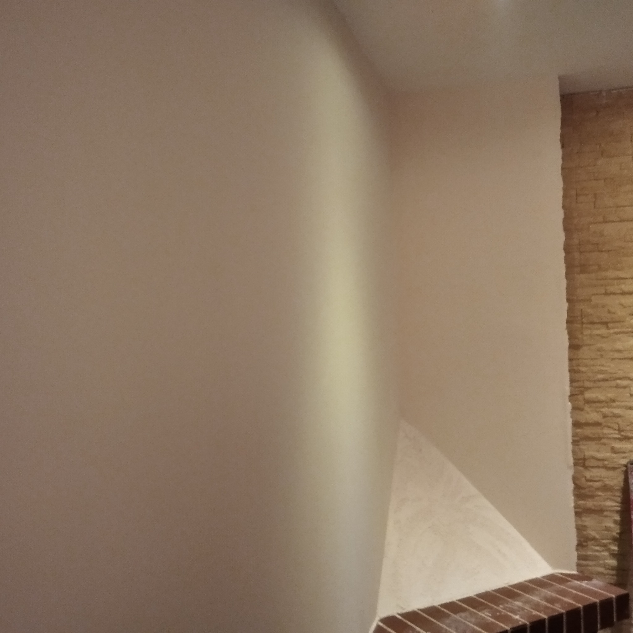 Gładka, beżowa ściana po tynkowaniu, częściowo pomalowana, z fragmentem ściany wykończonej dekoracyjną cegłą na krawędzi, widoczny cokół z brązowych płytek.