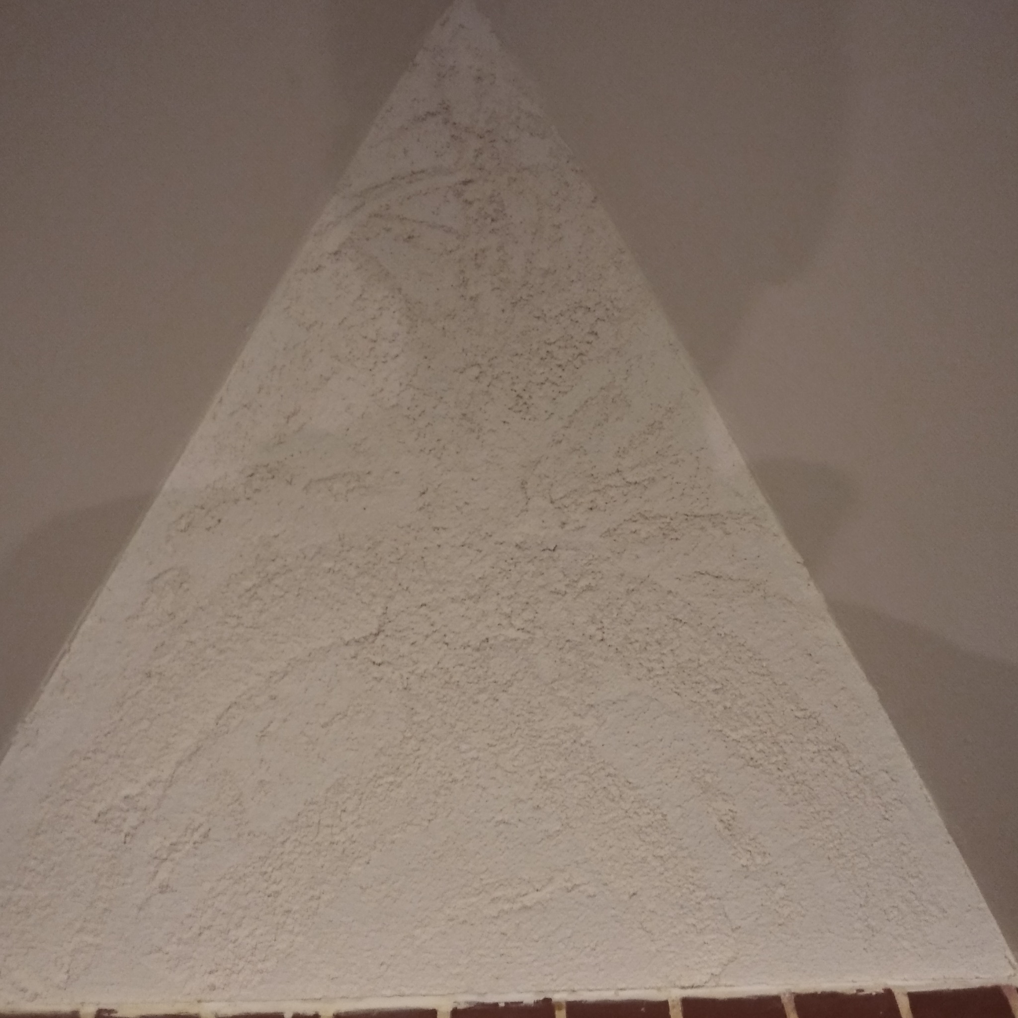 Trójkątna powierzchnia ściany z nałożonym tynkiem strukturalnym w odcieniach bieli, widoczna z bliska, z fragmentem ceglanej okładziny u podstawy.