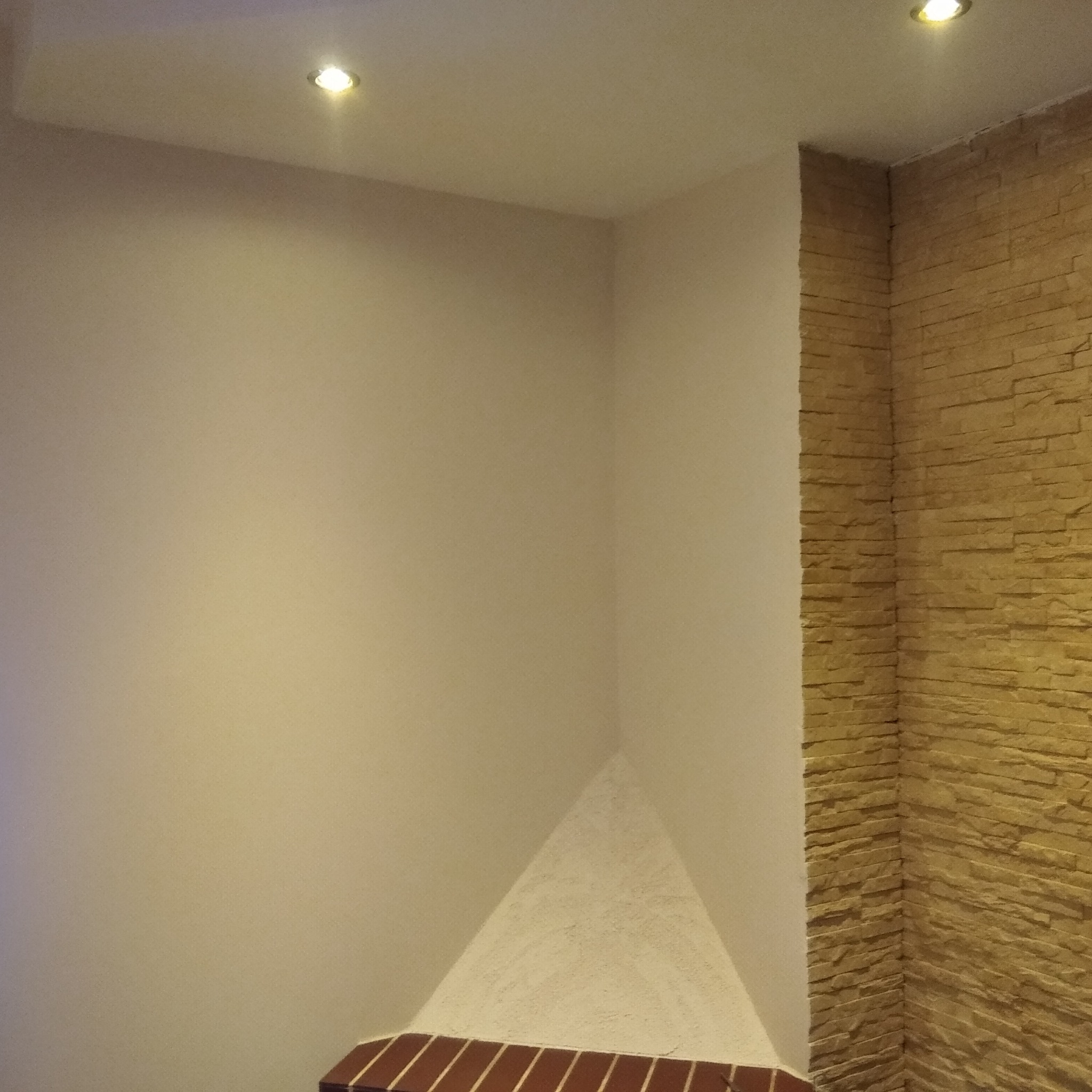 Ściana wewnętrzna z dekoracyjną okładziną z imitacji kamienia w odcieniach beżu, kontrastująca z gładką, jasną ścianą pomalowaną na kremowo. W suficie zamontowane dwa halogenowe punkty świetlne.