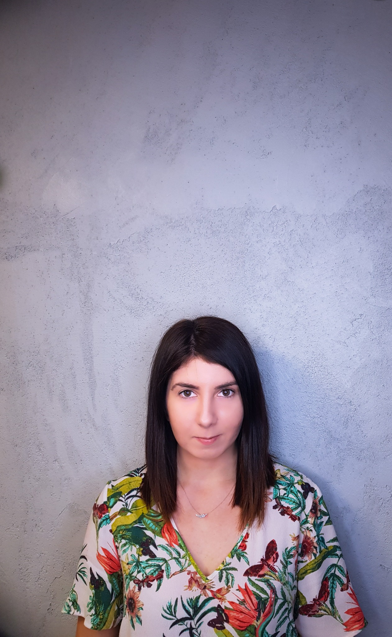 Portret kobiety z ciemnymi włosami w kwiecistej bluzce na tle szarej ściany z fakturą.