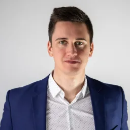 Portret młodego mężczyzny w niebieskiej marynarce i koszuli w kropki, z zielonymi oczami, na jasnym tle.