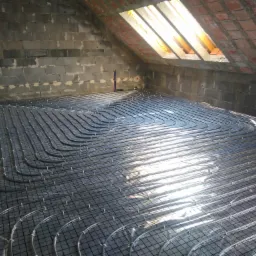 Instalacja ogrzewania podłogowego na poddaszu, rury ułożone na folii refleksyjnej, widoczne surowe ściany z pustaków i okno dachowe.