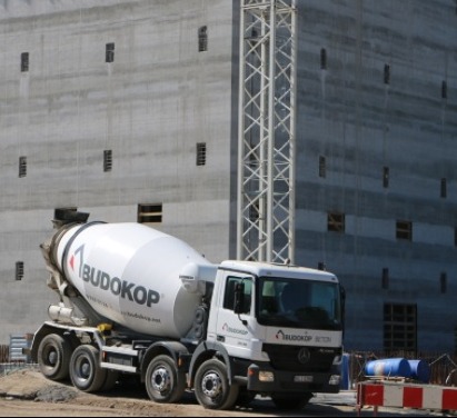 Ciężarówka z betonomieszarką marki Budokop na placu budowy, na tle surowego, betonowego budynku z małymi oknami i stalowej konstrukcji dźwigu.