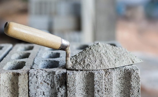 Kielnia z zaprawą murarską leży na betonowych pustakach z otworami, widoczne tło w odcieniach beżu i szarości.