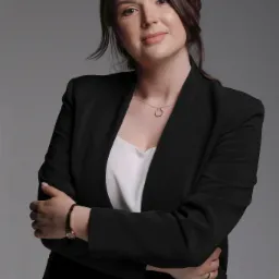 Portret uśmiechniętej kobiety w czarnej marynarce i białej bluzce, z rękami skrzyżowanymi na piersiach, na szarym tle.