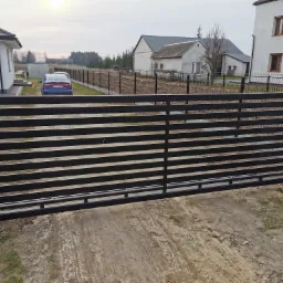 Nowoczesna, przesuwna brama wjazdowa z poziomych, czarnych profili, osadzona między szarymi, betonowymi słupkami, na tle zabudowy jednorodzinnej i zaoranego pola.