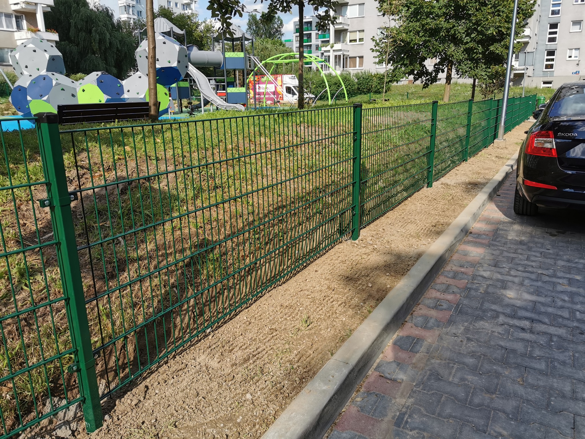 Nowo zamontowane, zielone ogrodzenie panelowe oddzielające plac zabaw dla dzieci od parkingu, widoczne z perspektywy chodnika z kostki brukowej, z czarnym samochodem Skoda w kadrze.