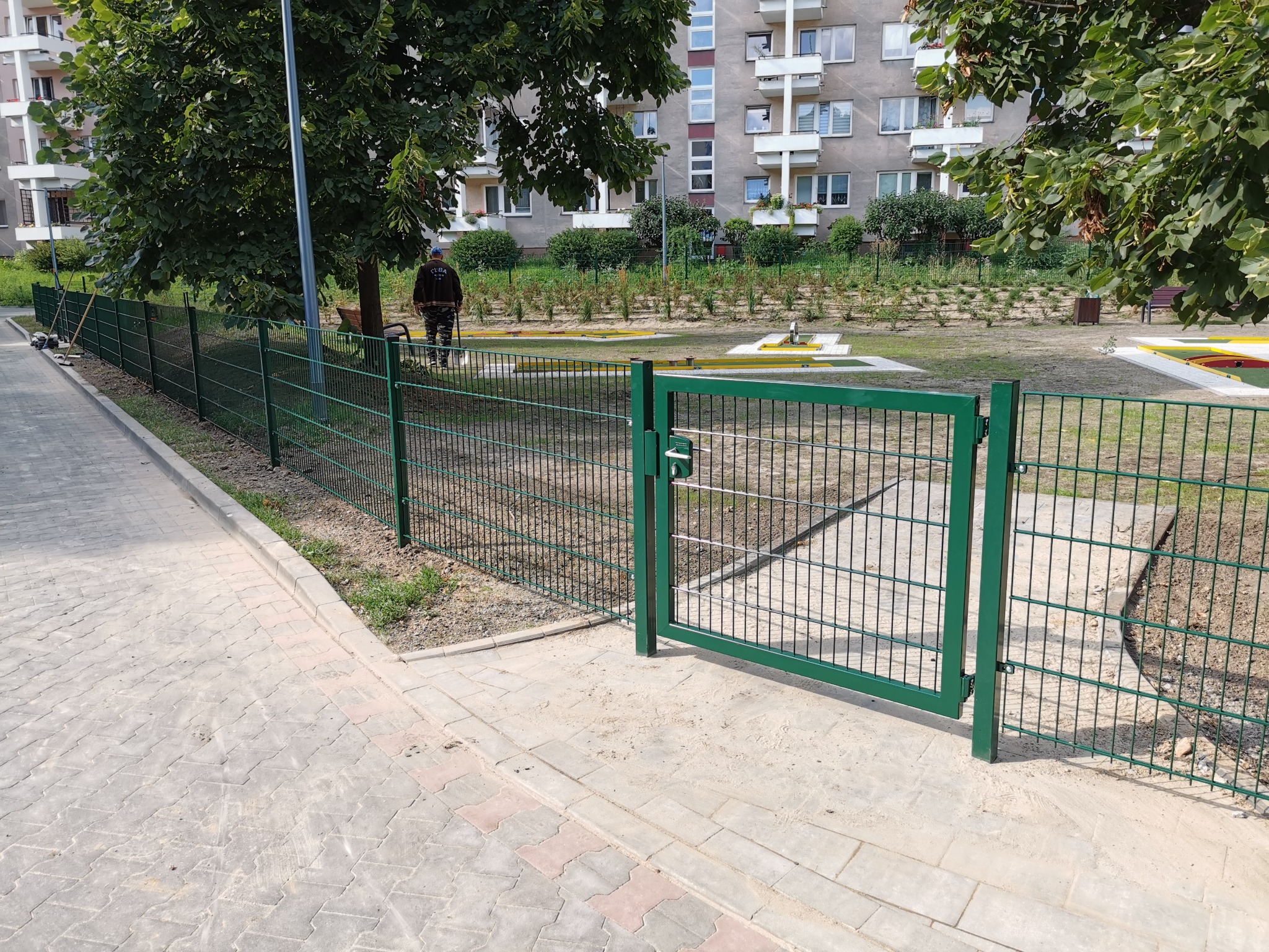 Otwarta, zielona furtka w siatkowym ogrodzeniu na tle osiedlowego placu zabaw i bloku mieszkalnego.