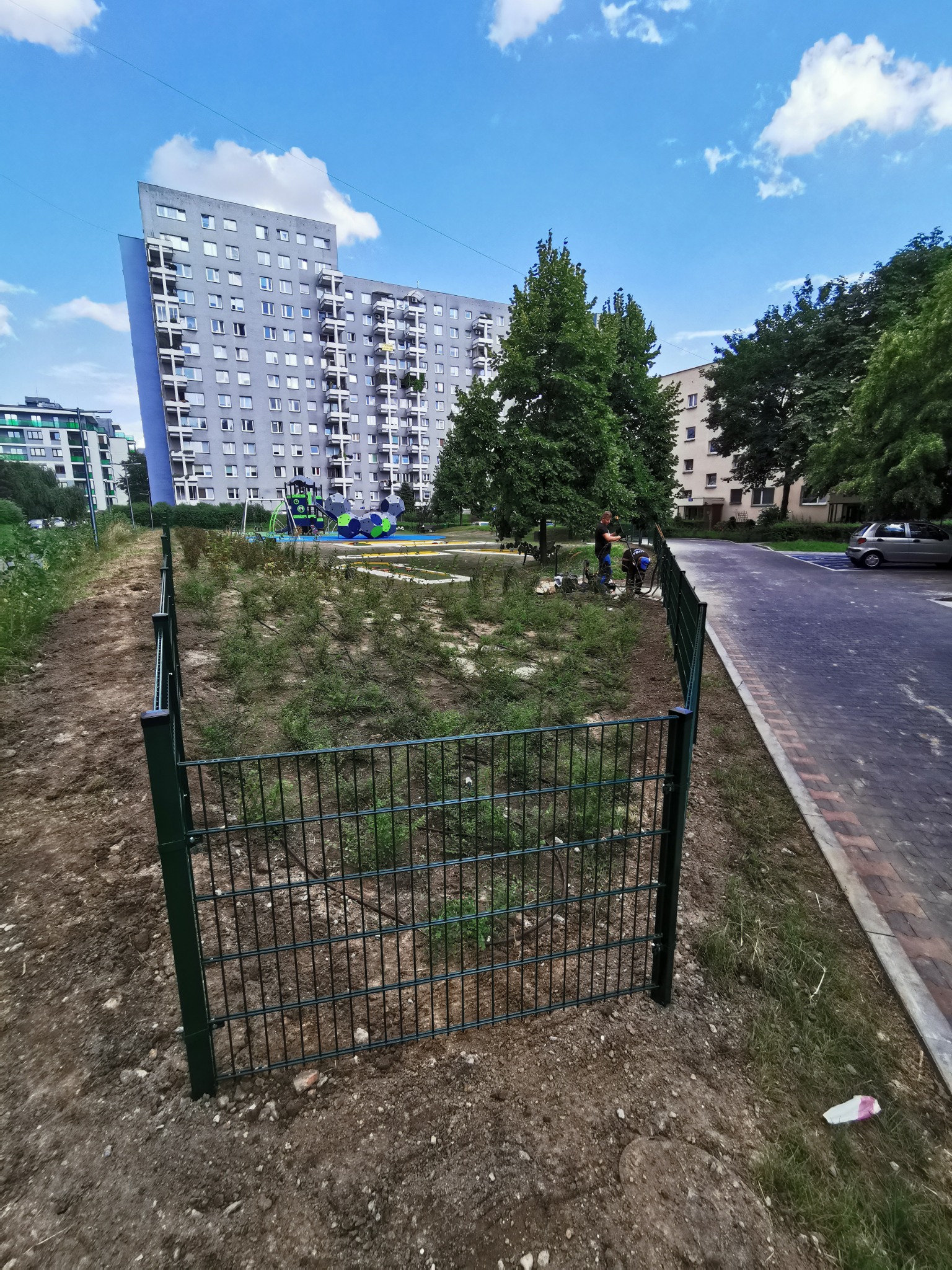 Fragment nowo zamontowanego, zielonego ogrodzenia panelowego na grządkach w otoczeniu bloków mieszkalnych, z widocznymi pracownikami w tle.