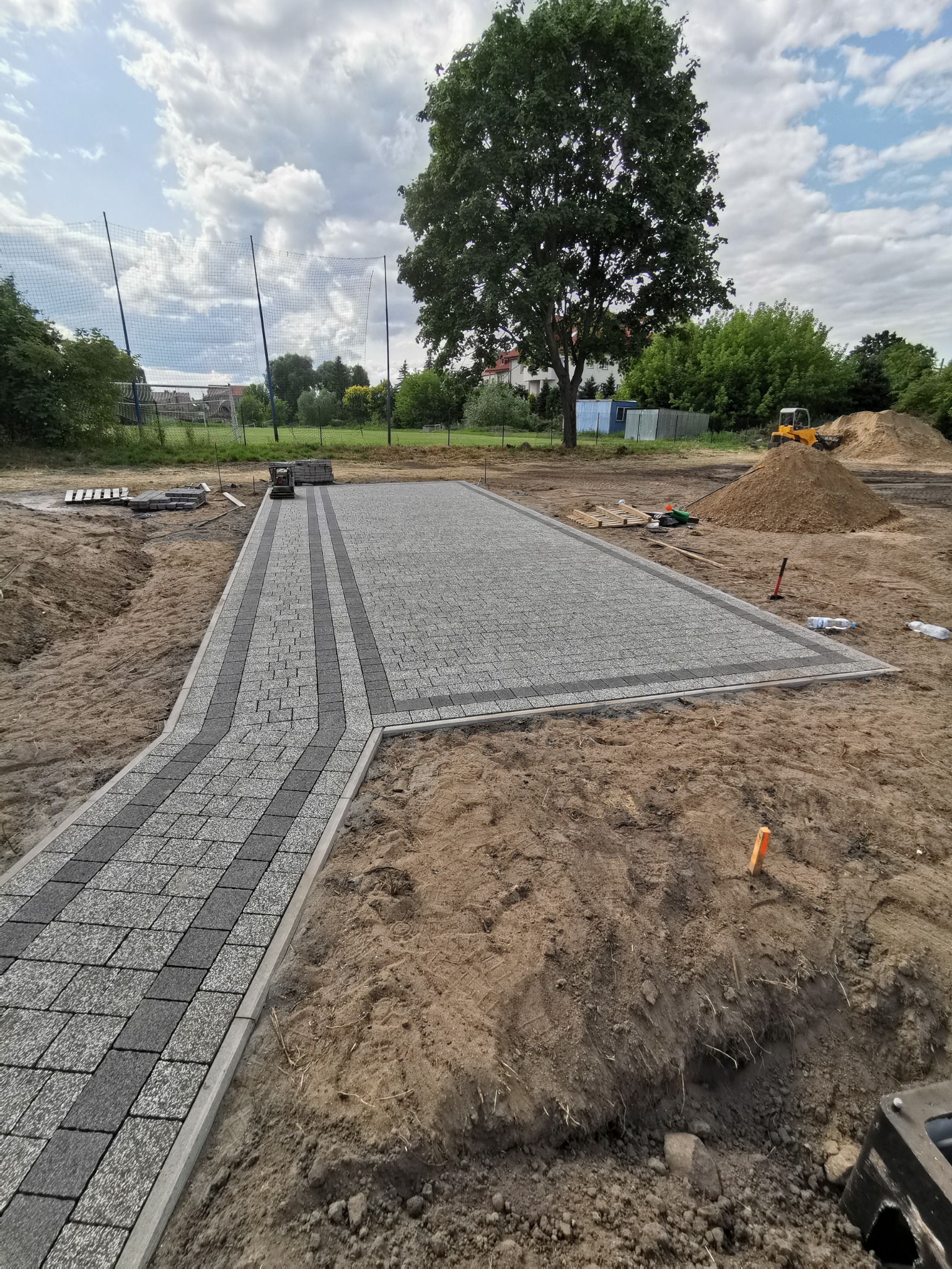Układanie kostki brukowej na posesji, widoczny fragment ścieżki i placu z jasnoszarej i ciemnoszarej kostki, przygotowany teren, w tle koparka i hałda piasku.