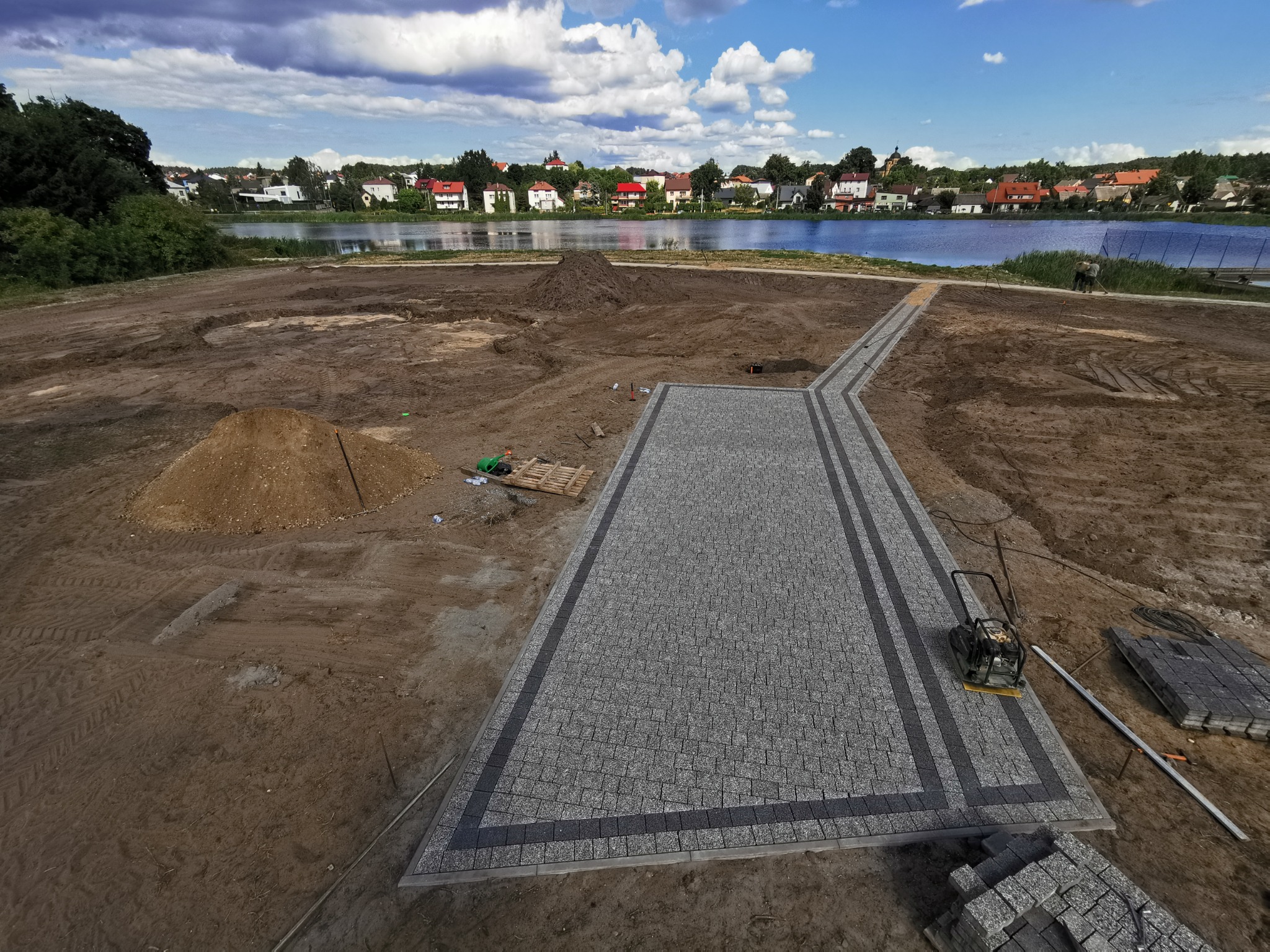 Układanie ścieżki z kostki brukowej w pobliżu jeziora, widoczny ubijak wibracyjny i palety z kostką, w tle zabudowa mieszkalna.