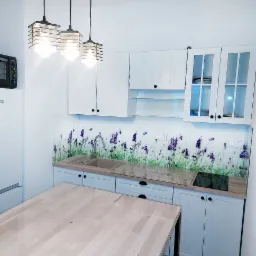 Jasna kuchnia z białymi szafkami, szklanym panelem z motywem lawendy oraz drewnianym blatem i stołem, oświetlona trzema wiszącymi lampami w stylu industrialnym.