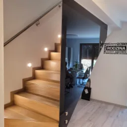 Drewniane schody z podświetleniem w ścianie i metalową poręczą, oddzielone od części salonowej szklaną ścianą z widocznym odbiciem, w tle stół z krzesłami i napis na ścianie: rodzina, szczęście...