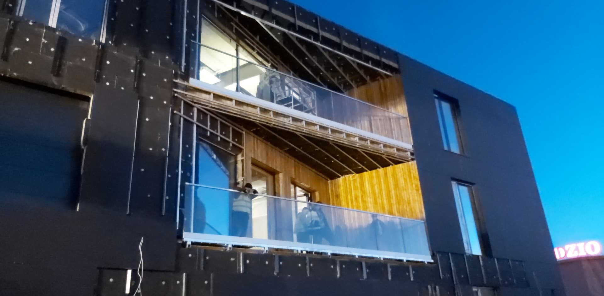 Montaż balustrad szklanych na dwóch balkonach budynku w trakcie budowy, widoczne elementy konstrukcyjne i pracownicy w kamizelkach odblaskowych.
