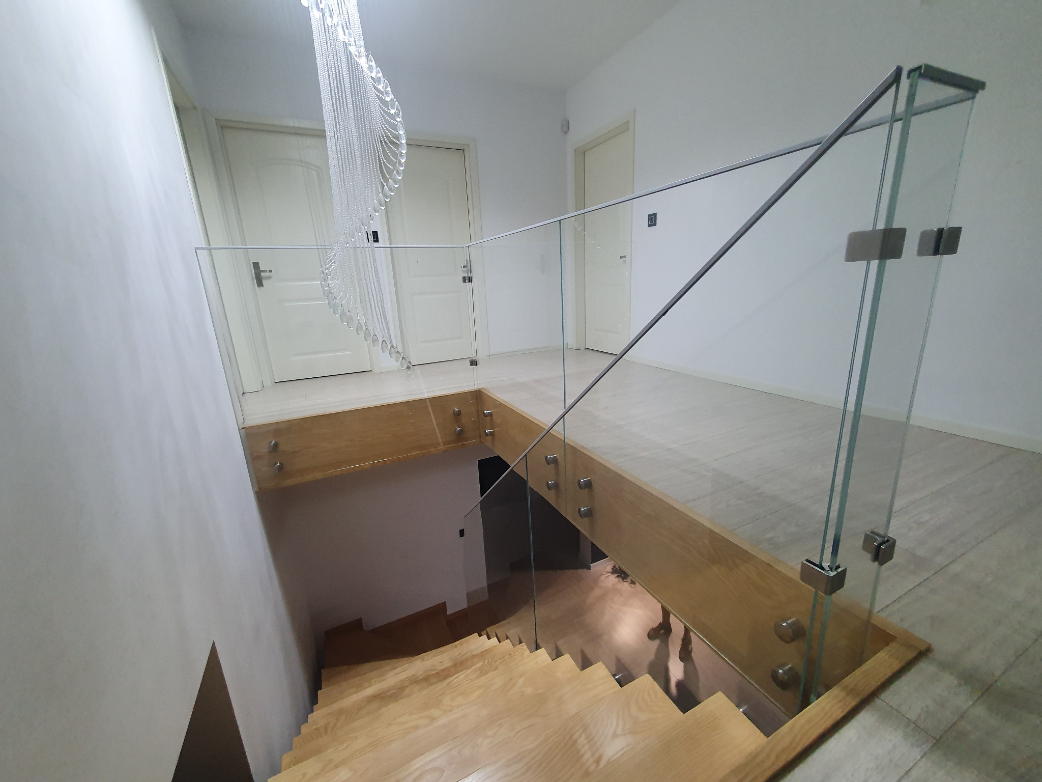 Wewnętrzna klatka schodowa z drewnianymi schodami, zabezpieczona szkłem mocowanym punktowo, widok z góry na dół, dekoracyjny żyrandol w tle