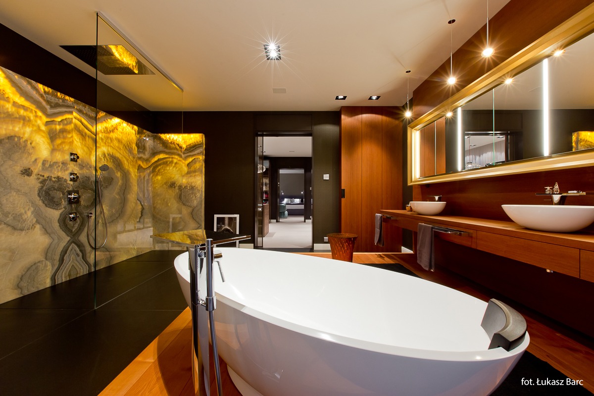 Luksusowa łazienka z wolnostojącą wanną, podświetlaną ścianą z kamienia onyksowego, drewnianymi szafkami i umywalkami nablatowymi.