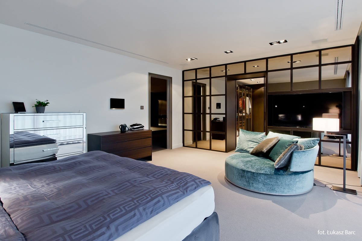 Przestronna sypialnia z łóżkiem z niebieską pościelą w geometryczny wzór, lustrzaną komodą, garderobą oddzieloną szklaną ścianą w czarnej ramie oraz okrągłą sofą w kolorze turkusowym.