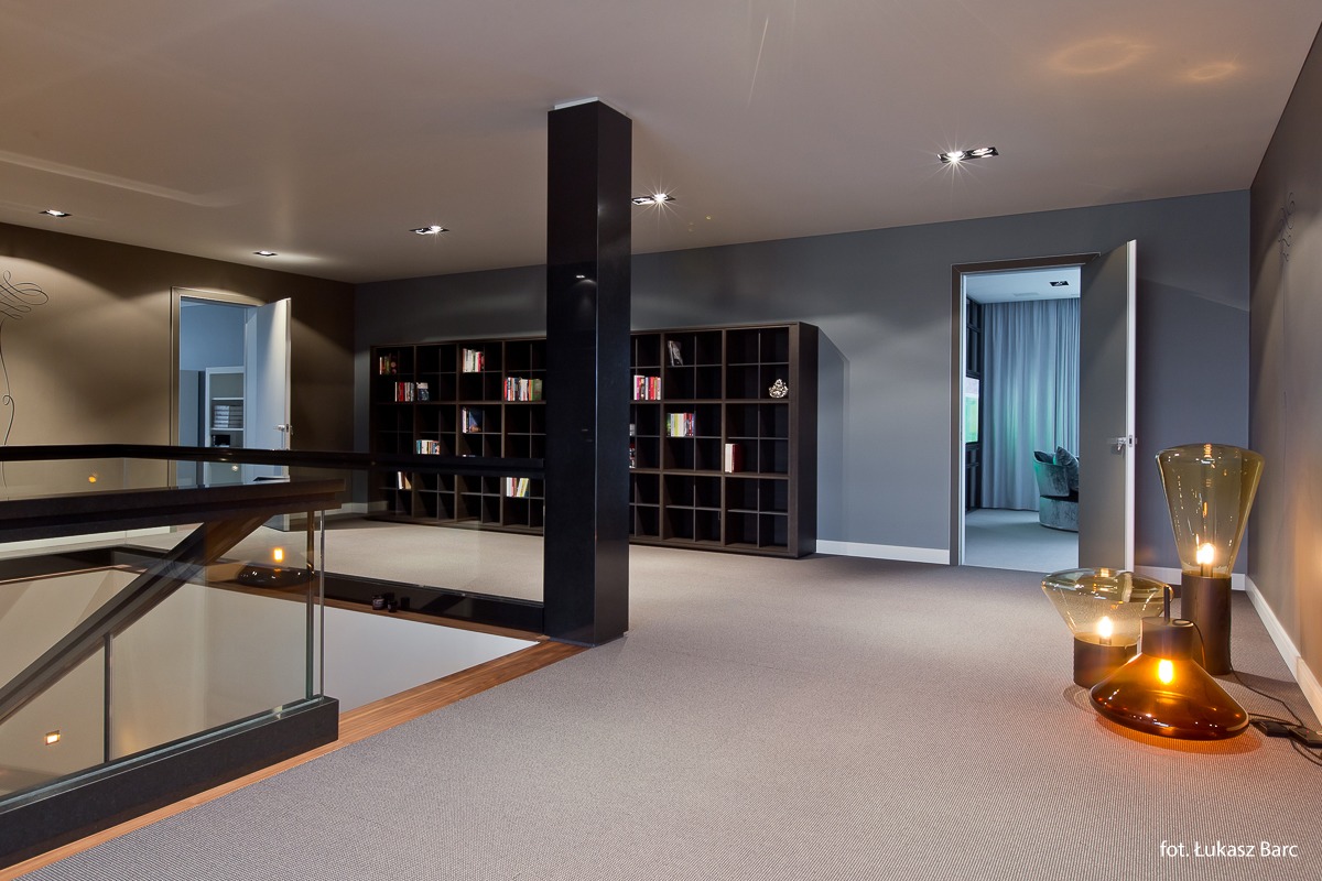 Przestronne wnętrze domu z antresolą, ciemnymi regałami na książki i dekoracyjnymi lampami podłogowymi, widok z góry na szklaną balustradę.