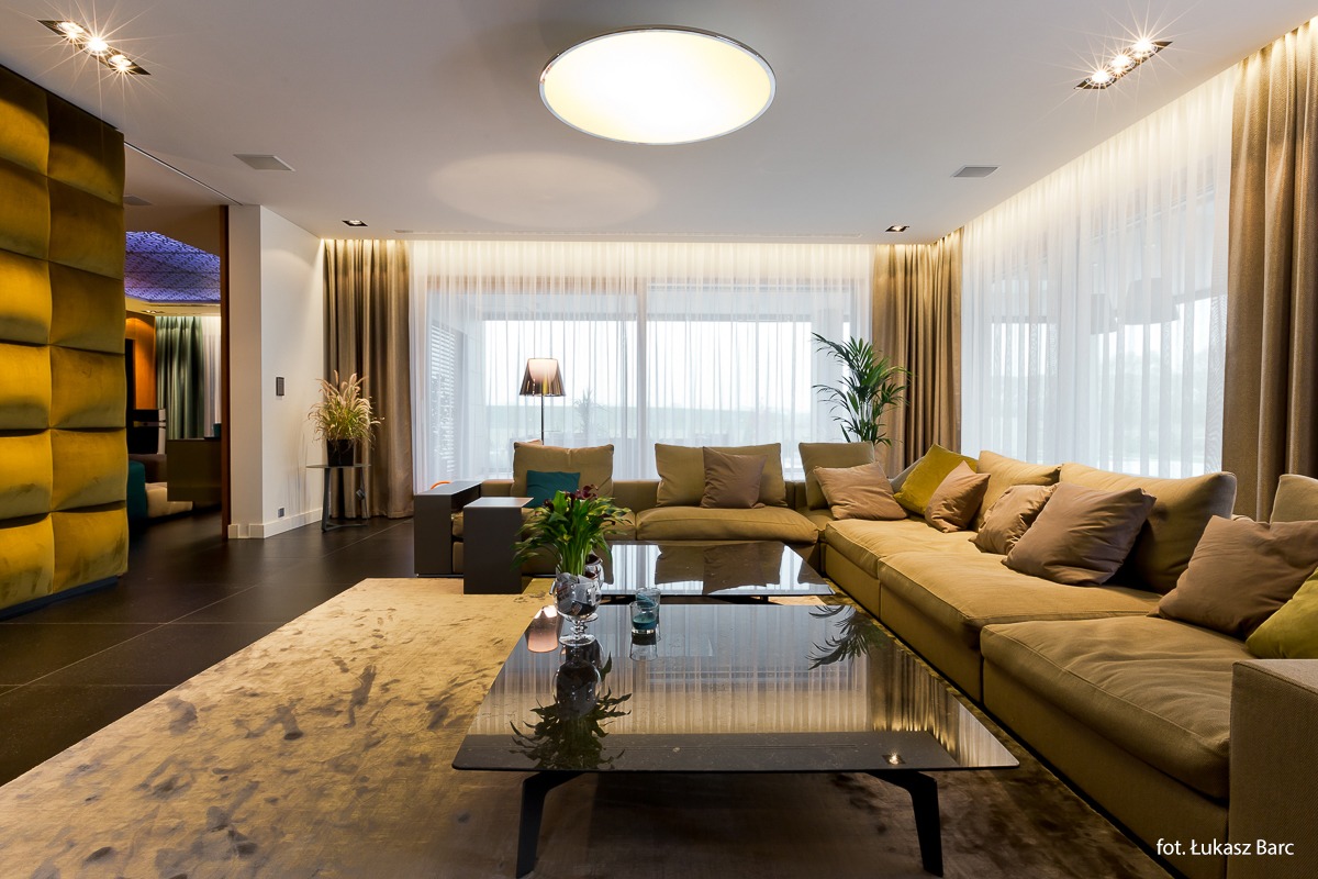 Przestronny salon z dużym narożnikiem w odcieniach beżu, szklanym stolikiem kawowym i subtelnym oświetleniem punktowym, widok na okno z zasłonami.