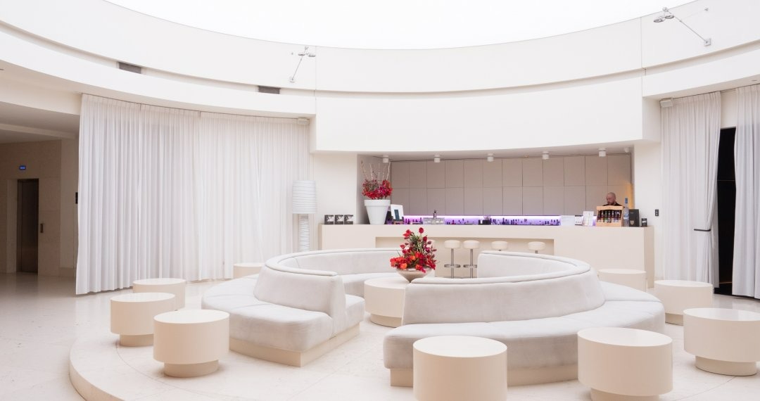 Jasne, okrągłe wnętrze z półkolistą sofą, okrągłymi stolikami, barem w tle i białymi zasłonami, oświetlone subtelnym światłem.