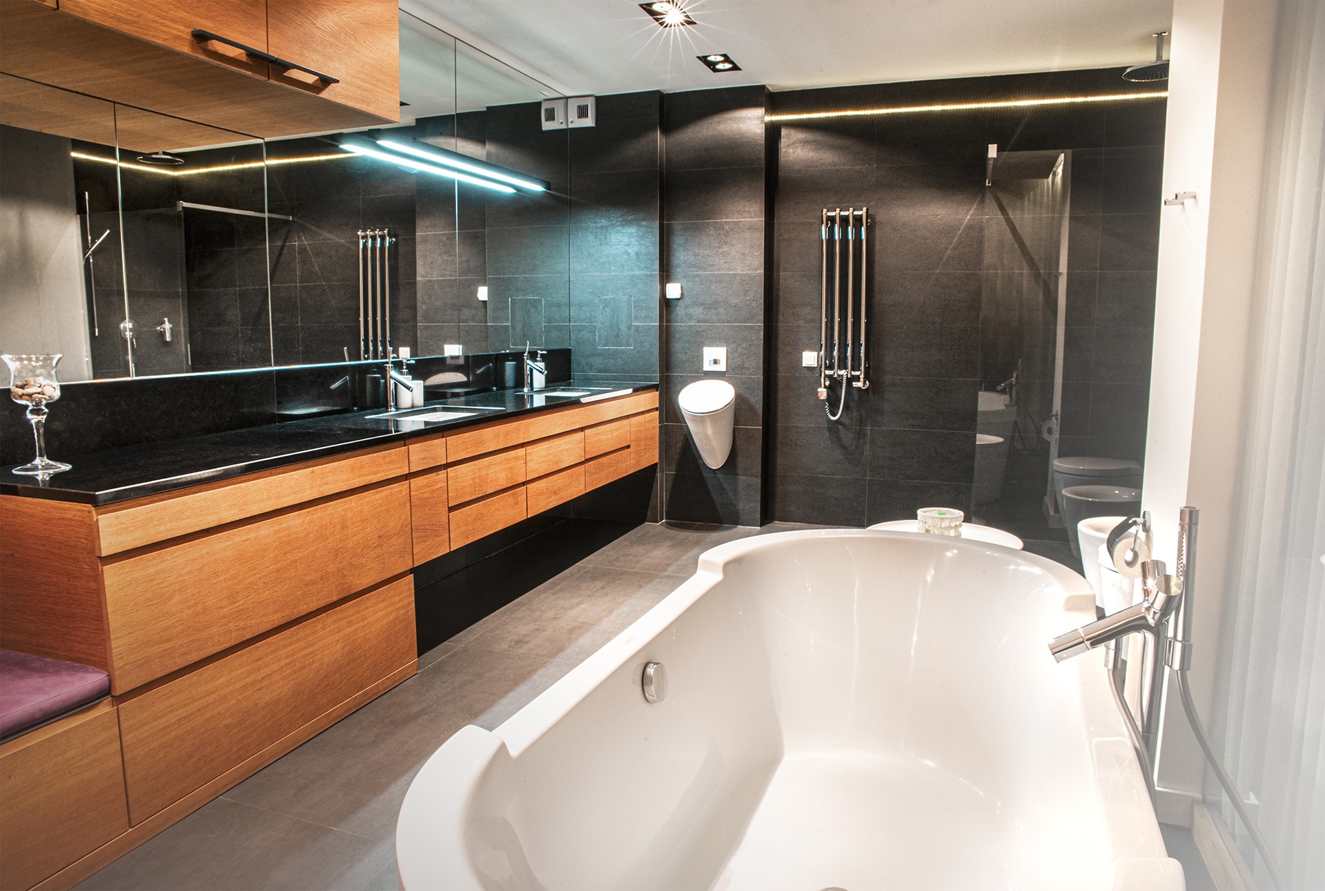 Nowoczesna łazienka z czarnymi płytkami, drewnianą zabudową umywalki z czarnym blatem, białą wanną wolnostojącą i podświetleniem LED.