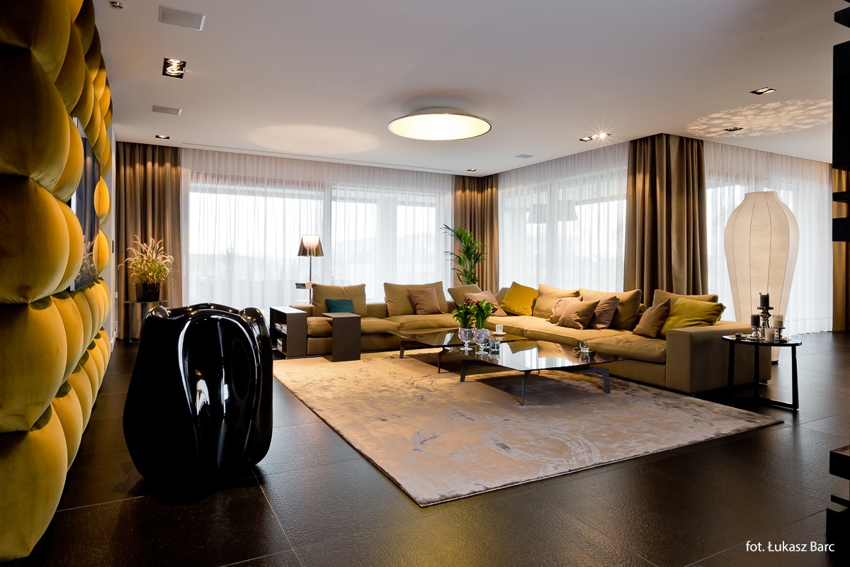Przestronny salon z narożną sofą w odcieniach beżu, szklanym stolikiem kawowym, dywanem z abstrakcyjnym wzorem i dekoracyjną ścianą z żółtymi panelami. Czarny, błyszczący fotel rzeźbiarski w lewym...