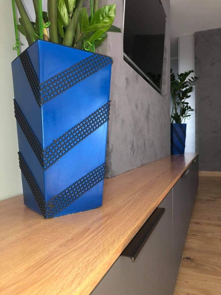 Minimalistyczne wnętrze z betonową ścianą, drewnianą szafką RTV i dwoma niebieskimi donicami o geometrycznym wzorze, w których rosną zielone rośliny.